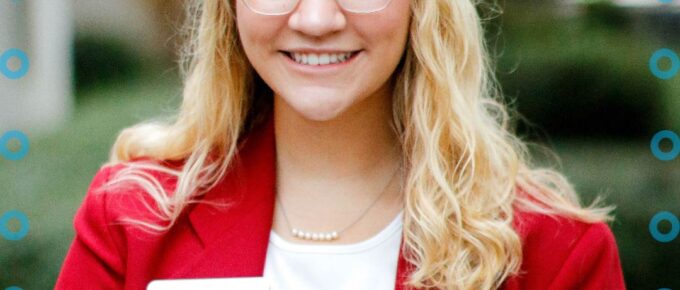 A young blond woman with glasses wearing a red jacket and a blurred name badge smiling towards the camera