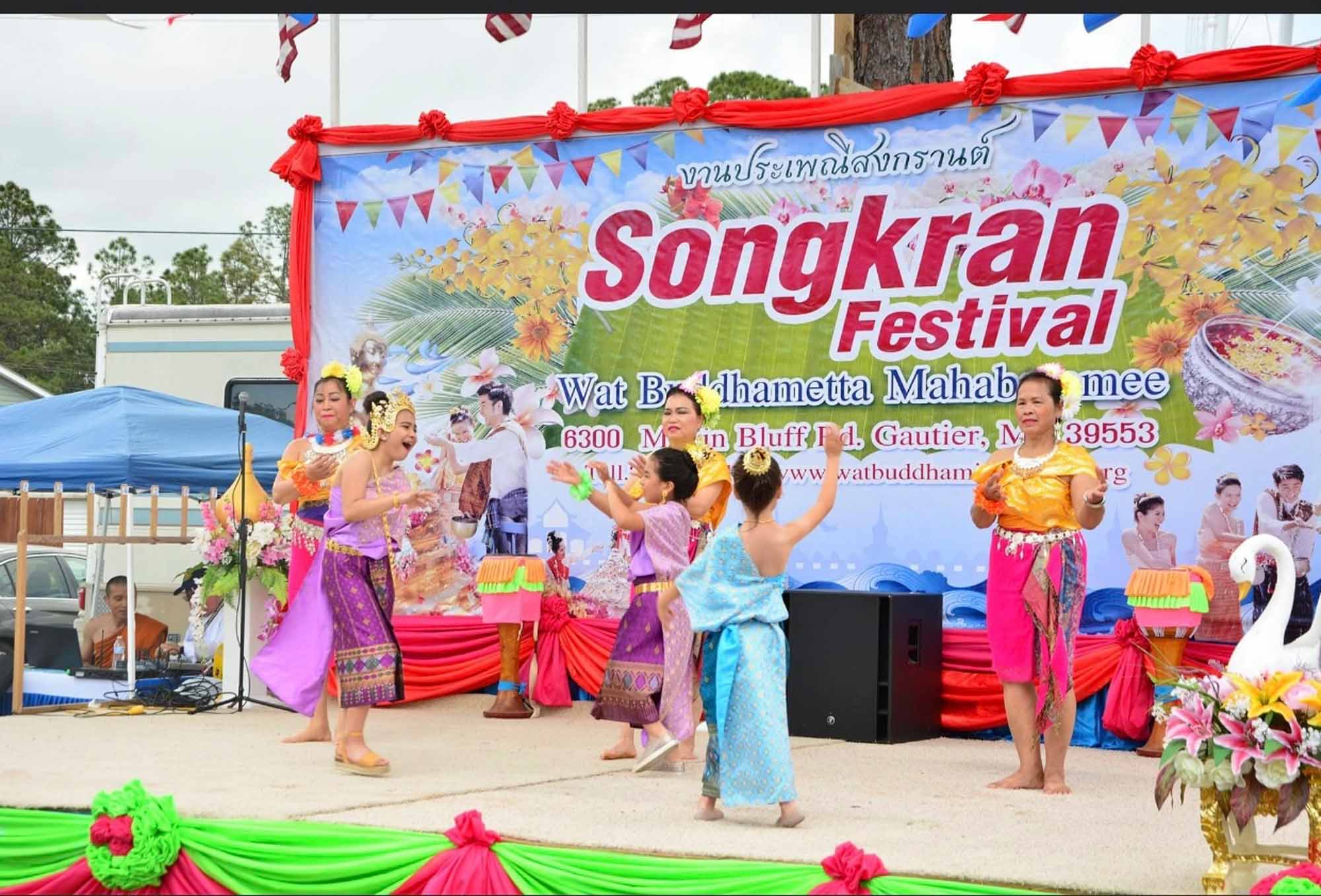 Children and adults participating in a festival on stage wearing colorful clothing and dancing. The sign in the background says Songkran Festival.