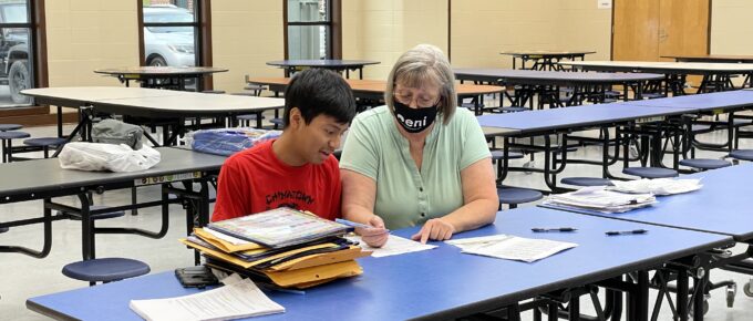 Blountsville Community Liaison Debra Goble sits with a young Hispanic teen at a school lunch table. Debra is wearing a mask with the ENI logo. There are several papers and folders in front of them. They are working on registering the teen for classes. Debra is writing while the teen is speaking.