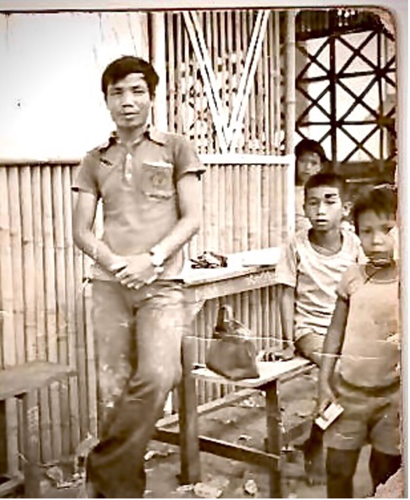 Lao man leans on a picnic table with his arms crossed. Three young Lao children appear next to him; one is standing, one is sitting on the bench, and one is peaking out from behind the wall behind the picnic table. The picture is sepia because it's dated.