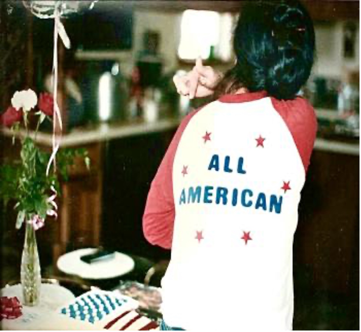 Lao woman stands with her back to the camera, pointing at the back of her t-shirt which reads "All American". Flowers, a balloon, and a cake with the American flag sit to her left.