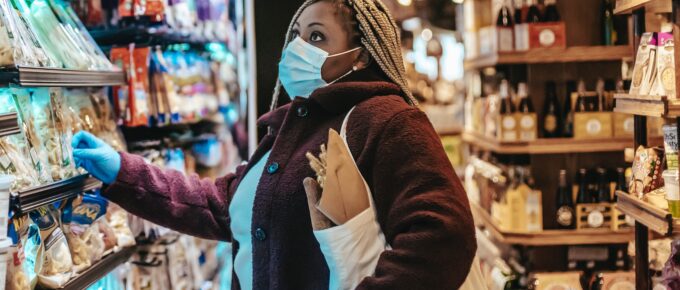 Black woman wearing surgical mask and gloves is holding a reusable bag filled with produce and looking up at the shelves in a grocery aisle