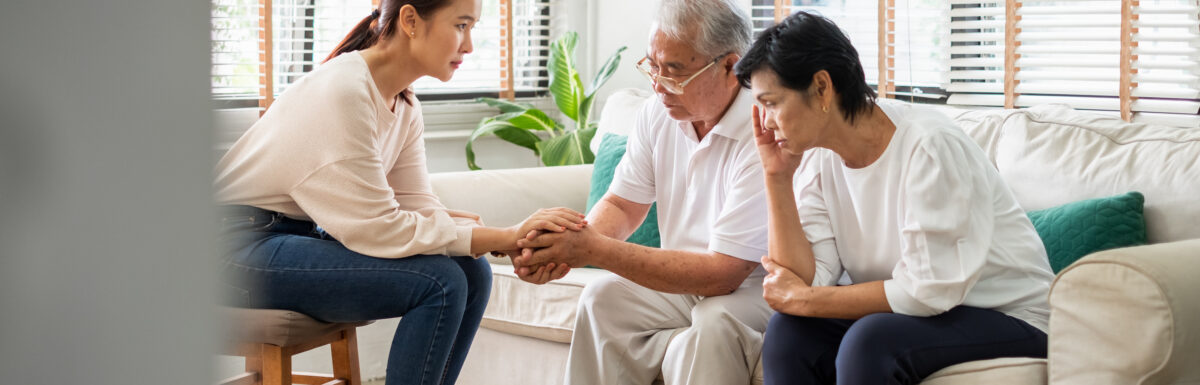 Female caregiver psychologist console Asian senior man and woman for mental health. Caregiver is holding hands of man while he sits on couch next to woman who is leaned over with her head in her hand.