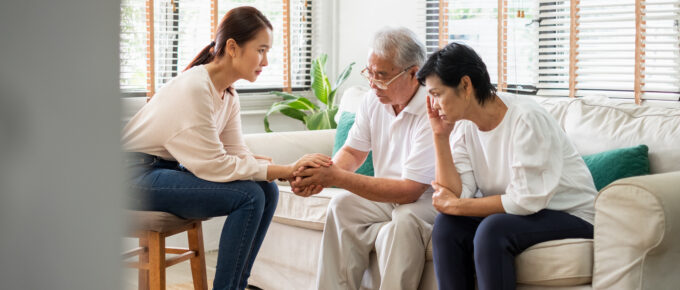 Female caregiver psychologist console Asian senior man and woman for mental health. Caregiver is holding hands of man while he sits on couch next to woman who is leaned over with her head in her hand.