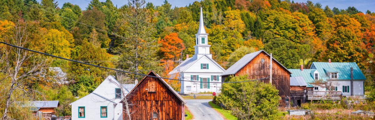 Landscape shot of a rural community with both white and wood-paneled rustic homes. In the middle sits a small white church with a tall steeple. The scenery is filled with autumn-colored trees and foliage.
