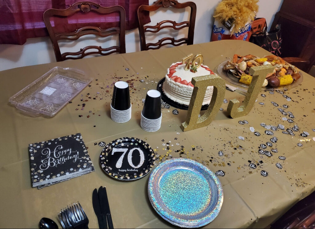 A dinner table decorated for a 70th birthday, including a cake, birthday plates and napkins, and confetti. A crawfish boil is plated to the side of the cake.