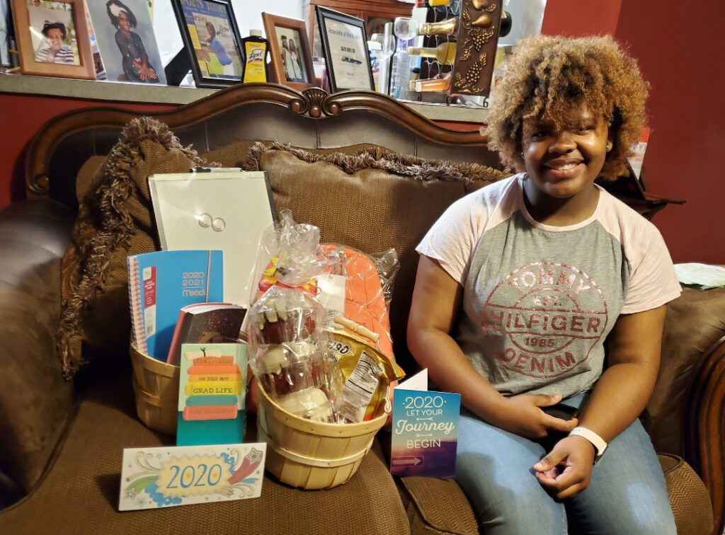 High school senior girl sits on a couch with gifts sitting next to her, including baskets filled with notebooks, snacks, a backpack, a small whiteboard, and three cards.