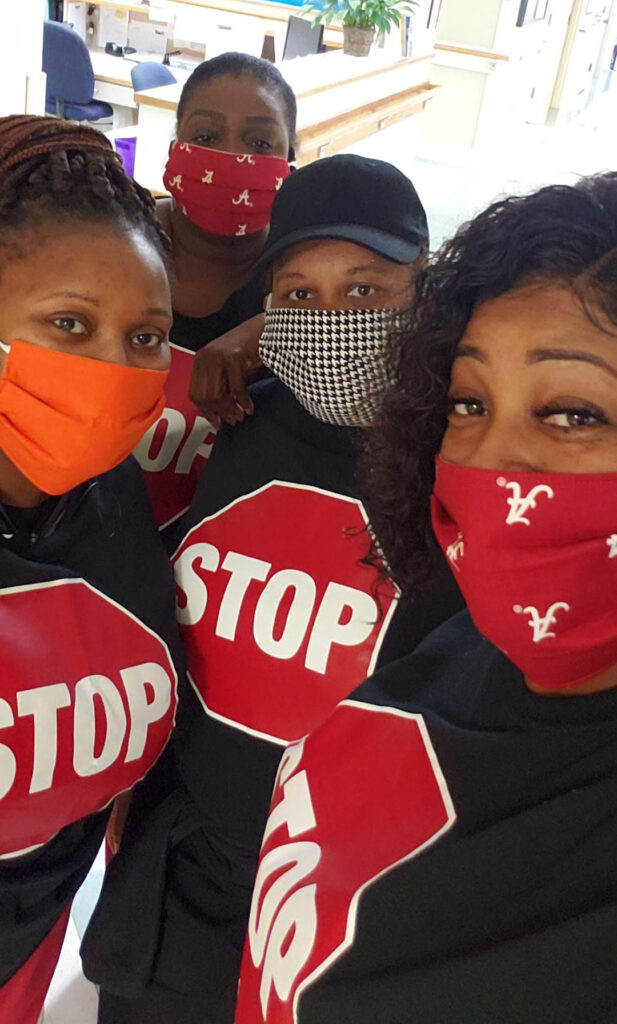 Four people take a selfie wearing masks and t-shirts that have a stop sign printed on the front. The background is a clinical setting.