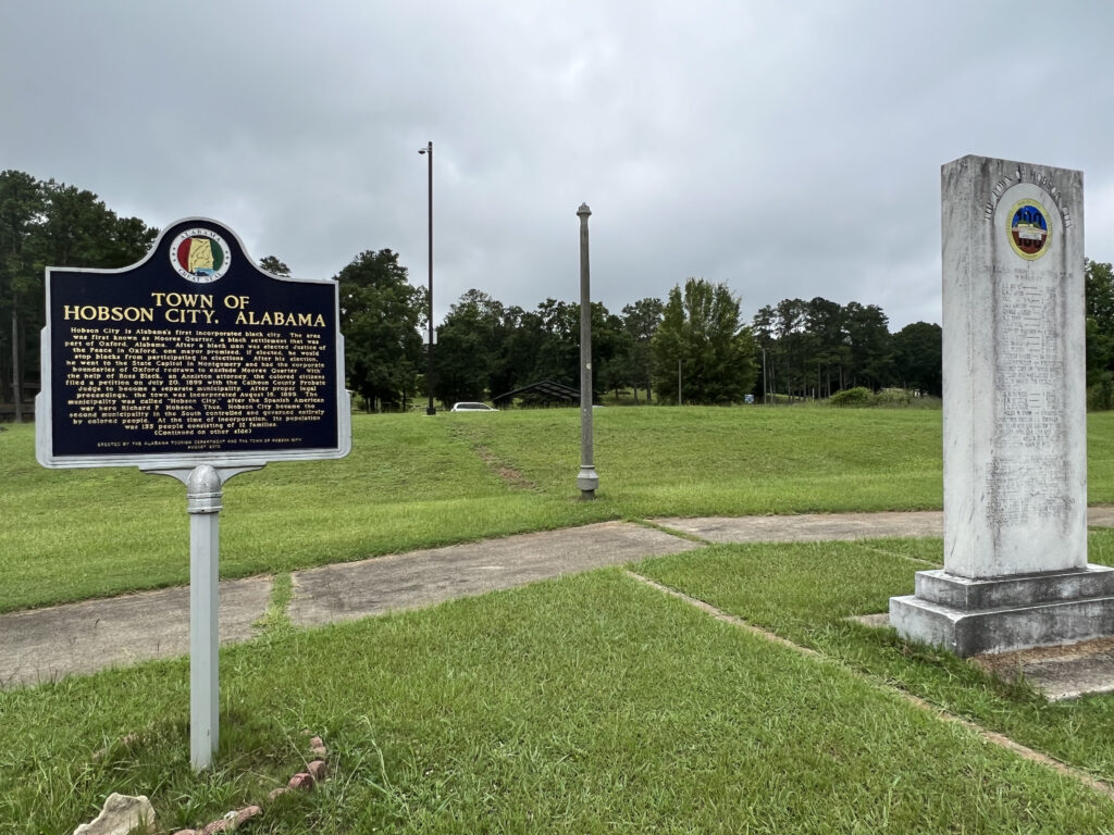 An Alabama historic marker featuring the "Town of Hobson City, Alabama" is situated in front of a grassy park with a walking path behind it. A few feet from the sign is a cement marker honoring Hobson City's centennial.
