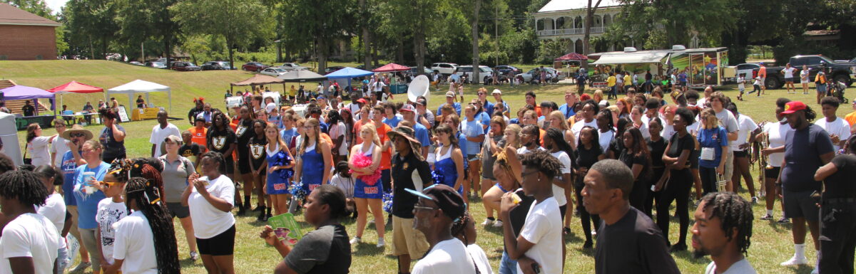 Hundreds of young people and families gather in summer clothes in a field next to the school. Some of the students are in band and cheerleading gear after just playing in a united band with multiple schools attending. In the background are tents with information tables and a food truck.