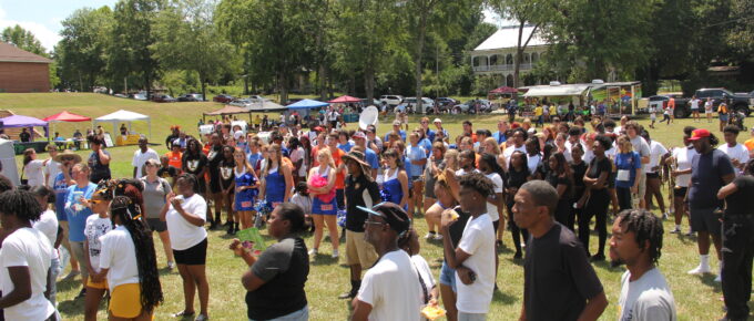 Hundreds of young people and families gather in summer clothes in a field next to the school. Some of the students are in band and cheerleading gear after just playing in a united band with multiple schools attending. In the background are tents with information tables and a food truck.