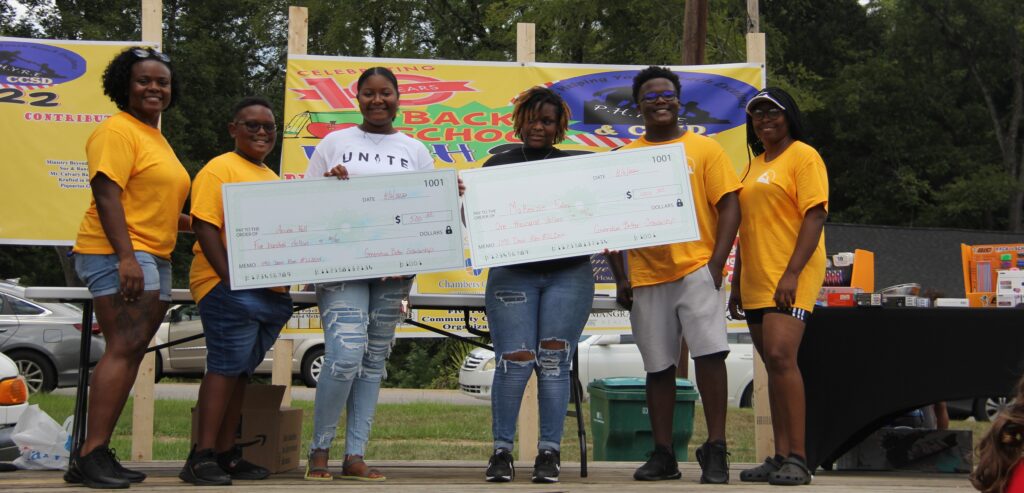 LaFayette Community Liaison Adrian Holloway and other P.H.Y.R.E. members pose with recipients of the Generation Better Scholarship on top of a stage. The two recipients are holding jumbo checks for $500 each.