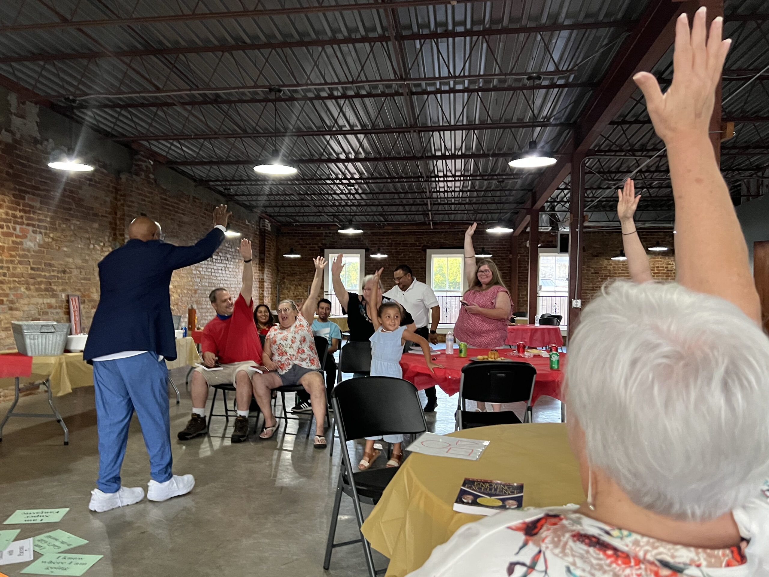 At the Collinsville ENI Kickoff meeting held on the top level of the library, motivational speaker Dr. Earl Suttle engages with the community members. Everyone pictured is raising a hand, laughing as Dr. Suttle asks them a question.