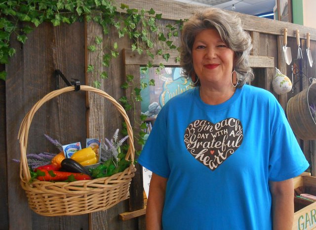 Jennifer Wilkins smiles in front of a display at the library. She is smiling and wearing a shirt that says "Begin each day with a grateful heart." The display behind her looks like a home with garden tools hanging. Next to her is a hanging basket filled with plastic fruit and flowers.