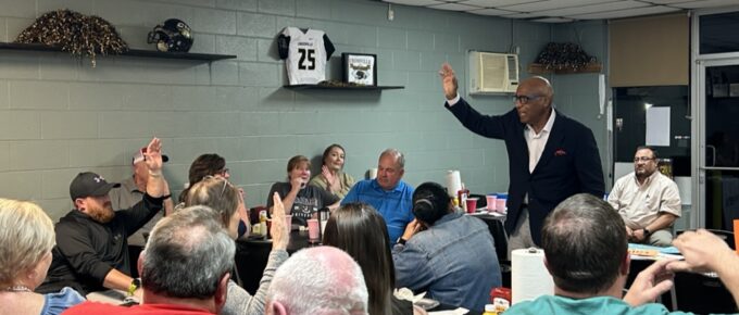 Dr. Earl Suttle stands in a group of people raising his hand, speaking. Several people in the group are also raising their hand. Dr Suttle is leading an exercise at the community kickoff for Crossville and Kilpatrick