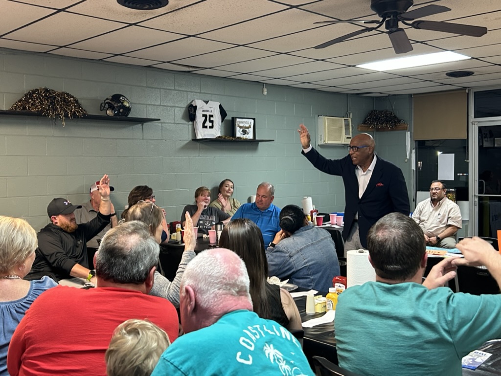 Dr. Earl Suttle stands in a group of people raising his hand, speaking. Several people in the group are also raising their hand. Dr Suttle is leading an exercise at the community kickoff for Crossville and Kilpatrick