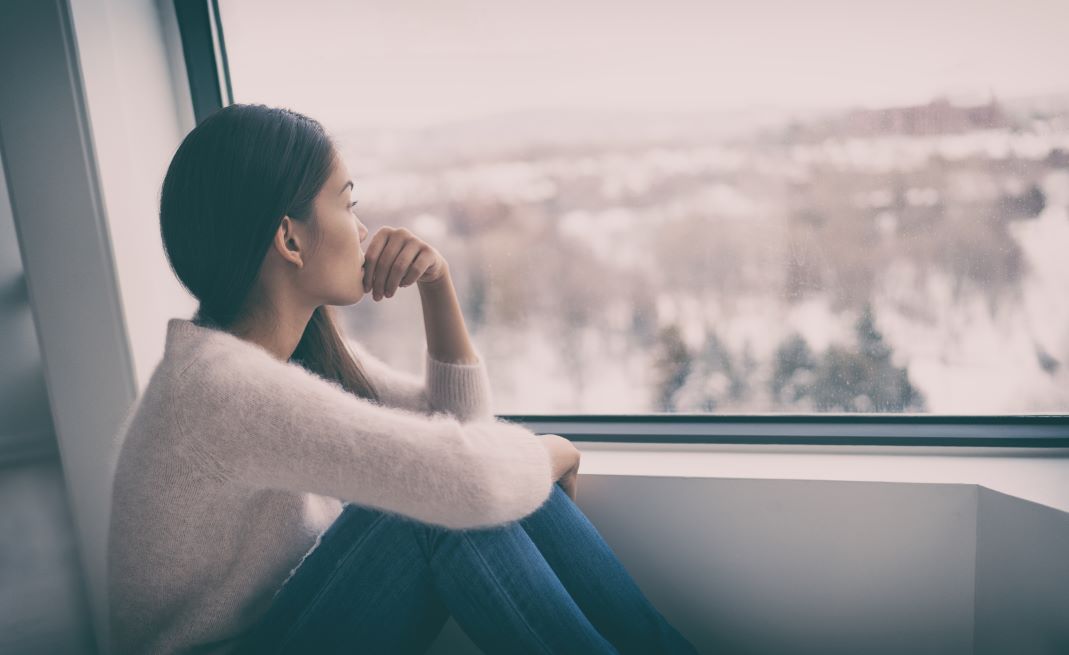 Young Asian woman sits with her knees pulled to her chest looking out a window