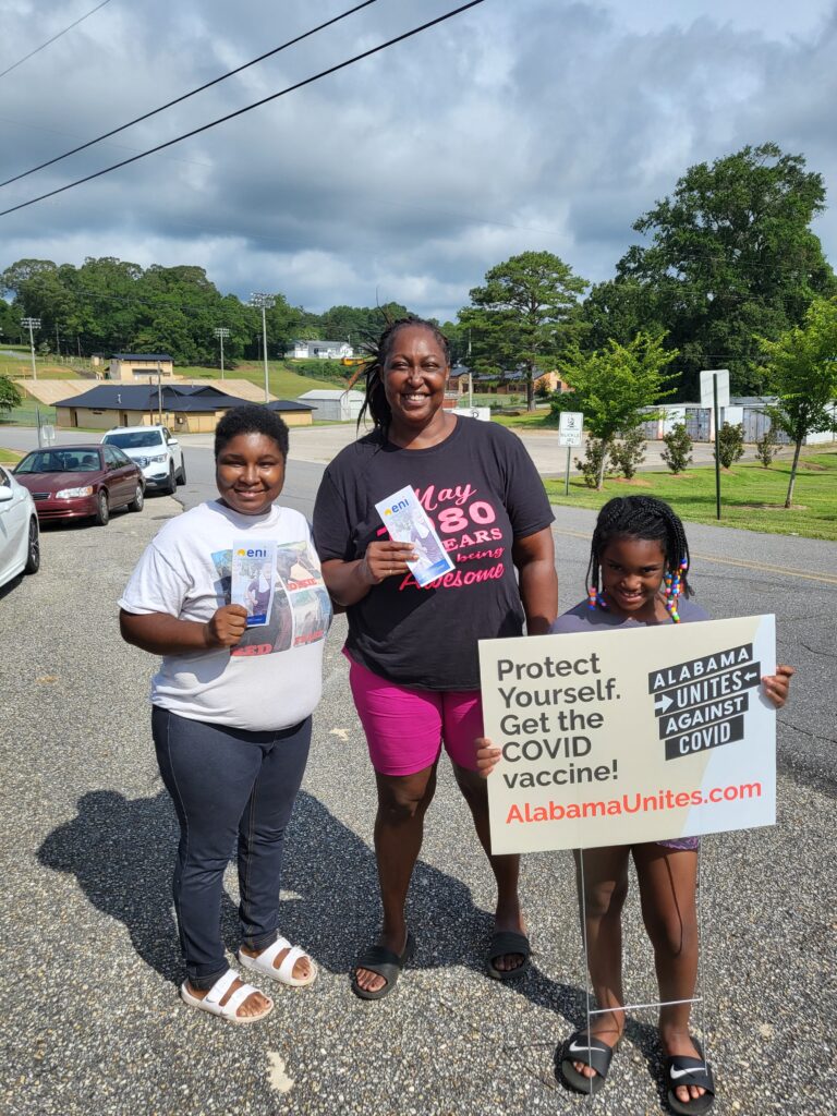 A woman with three children stand in the parking lot of an event. The woman and older child both hold an ENI brochure. The smaller child holds a sign from Alabama Unites Against COVID that reads "Protect Yourself. Get the COVID vaccine! AlabamaUnites.com"