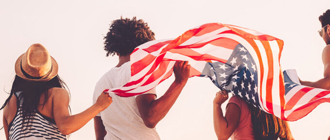 Four young adults stand in summer clothes and wave an American flag behind them