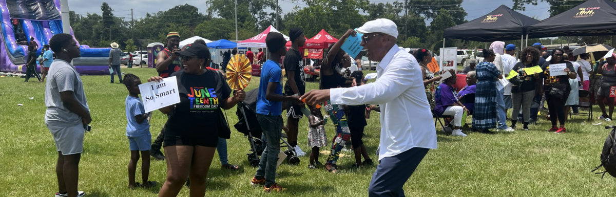 At the Juneteenth ENI kickoff in Prichard, Dr. Earl Suttle is laughing as he talks to a Prichard community member. The community member is wearing a Juneteenth Freedom Day t-shirt and holding a sign that says "I am so smart," part of an exercise Dr. Suttle does to engage with communities. In the background there is a bouncy slide, tents with tables, and lots of people. Some people in the background are holding other signs part of the same exercise.