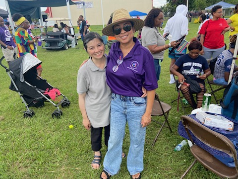 Kim-Lien Tran and Stephanie Khat pose at the Prichard Juneteenth event. There are many people in the background sitting on chairs or standing. Stephanie is wearing a t-shirt from the Alabama Department of Public Health.