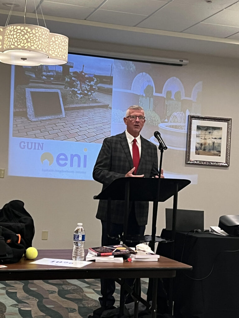 Keith Rhudy stands at a podium with a microphone. Behind him is a presentation with images of a park. The presentation is titled "Guin ENI"