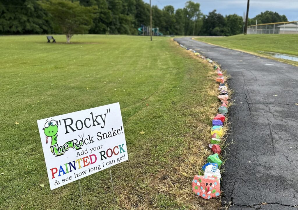 A sign is displayed in the grass next to a paved walking path. The sign has a cartoon graphic of a snake and reads "Rocky the Rock Snake! Add your Painted Rock and see how long I can get!" Dozens of painted rocks sit along the path; the front rock is painted to look like a snake head.