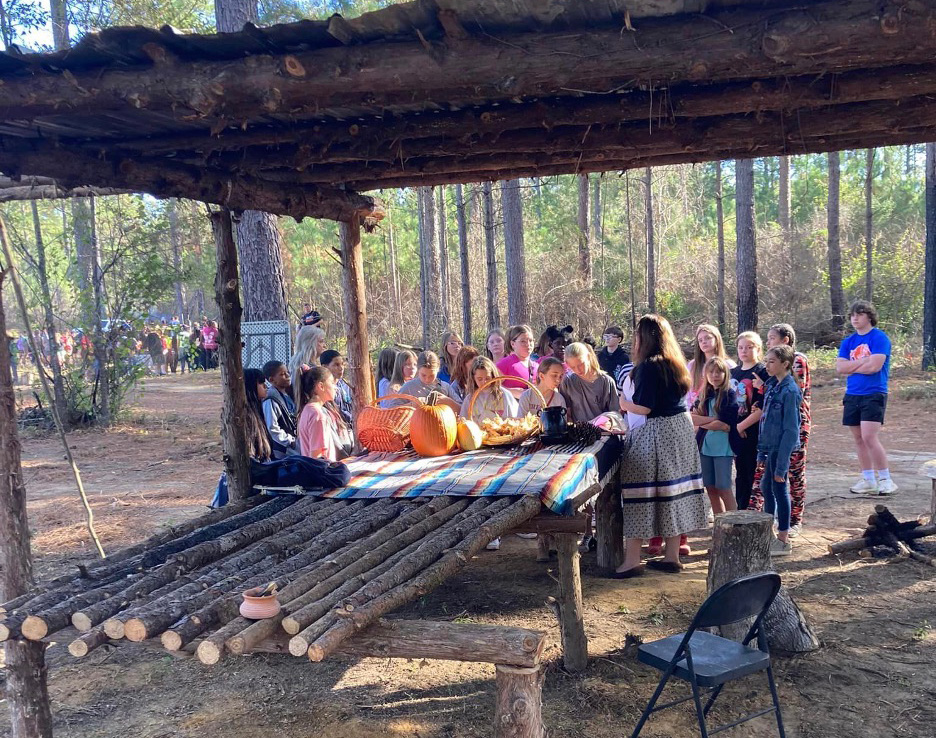 A group of children gather around an adult in traditional Ma-Chis dress with pumpkins and traditional foods on a table made of logs. The group stands under a shelter made of logs.