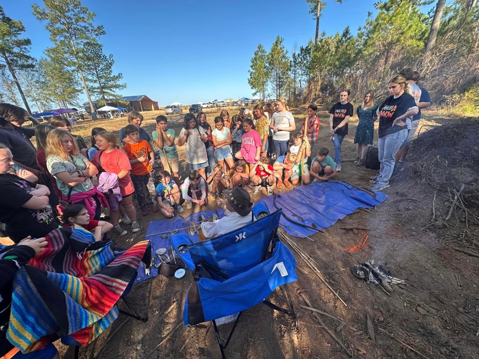 A group of people gathering around adults with various materials from the earth sitting on a tarp at their feet. There is a small fire with a small plume of smoke next to the group.