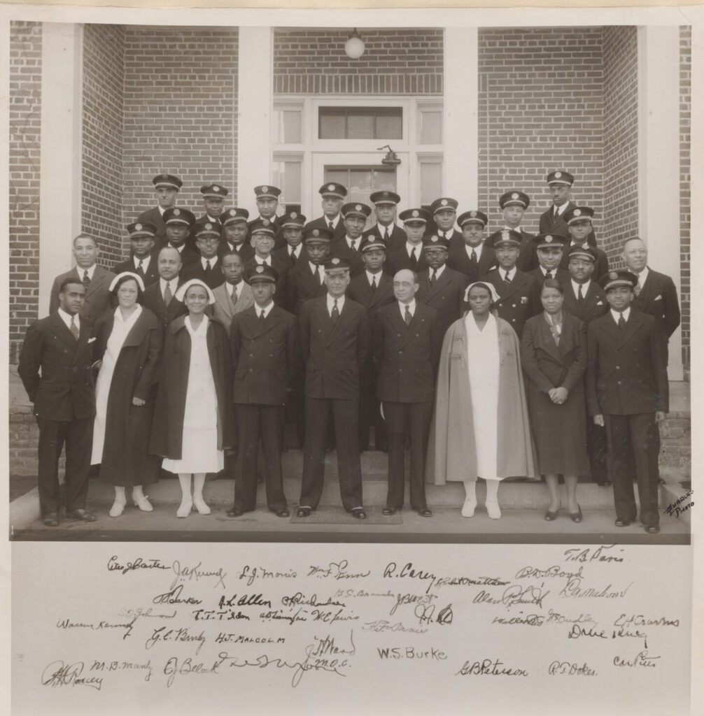 A group of Black soldiers and nurses pose in front of a brick building. The image is historic and in black and white.