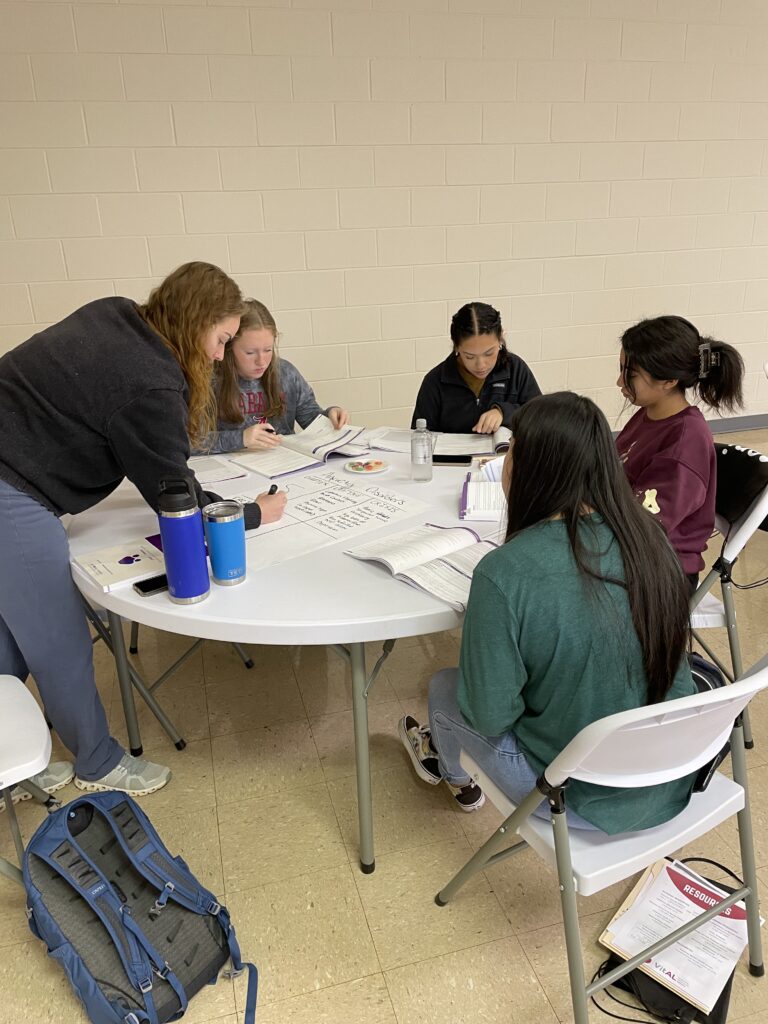 Five high school girls sit at a table, looking at workbooks and writing on a large sticky pad.